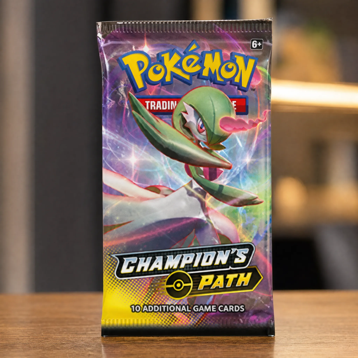 A Pokémon Trading Card Game booster pack for Champion's Path, featuring a green and white bird-like character on a colorful purple and pink background, standing on a wooden surface with a blurred indoor background.